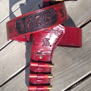 K&ouml;nnte beinhalten: Ein rotes Leder-G&uuml;rtel- und Pistolenholster-Set mit dem Text "K 1875 S" auf dem G&uuml;rtel. Das Holster hat ein Stacheldraht-Design und h&auml;lt mehrere rote Schrotpatronen. Ein h&ouml;lzerner Pistolen-Griff ist sichtbar.