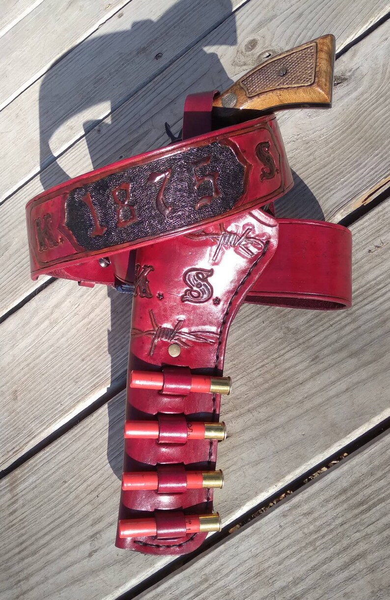 K&ouml;nnte beinhalten: Ein rotes Leder-G&uuml;rtel- und Pistolenholster-Set mit dem Text "K 1875 S" auf dem G&uuml;rtel. Das Holster hat ein Stacheldraht-Design und h&auml;lt mehrere rote Schrotpatronen. Ein h&ouml;lzerner Pistolen-Griff ist sichtbar.