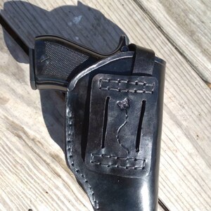 K&ouml;nnte beinhalten: Eine schwarze Lederholster mit einer Pistole darin. Die Holster hat ein dekoratives Schmetterlingsdesign und ist an den Kanten gen&auml;ht. Die Pistole ist schwarz mit einem strukturierten Griff.