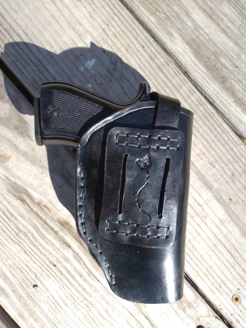 K&ouml;nnte beinhalten: Eine schwarze Lederholster mit einer Pistole darin. Die Holster hat ein dekoratives Schmetterlingsdesign und ist an den Kanten gen&auml;ht. Die Pistole ist schwarz mit einem strukturierten Griff.