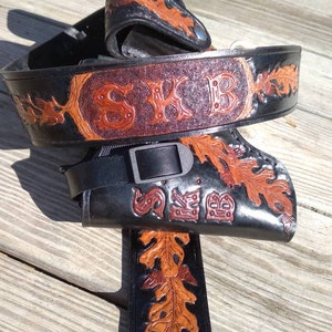 May include: A black leather belt and holster set with brown oak leaf and scrollwork designs. The belt features the initials "SKB" in a decorative font. The set is made of thick leather and has a buckle closure.