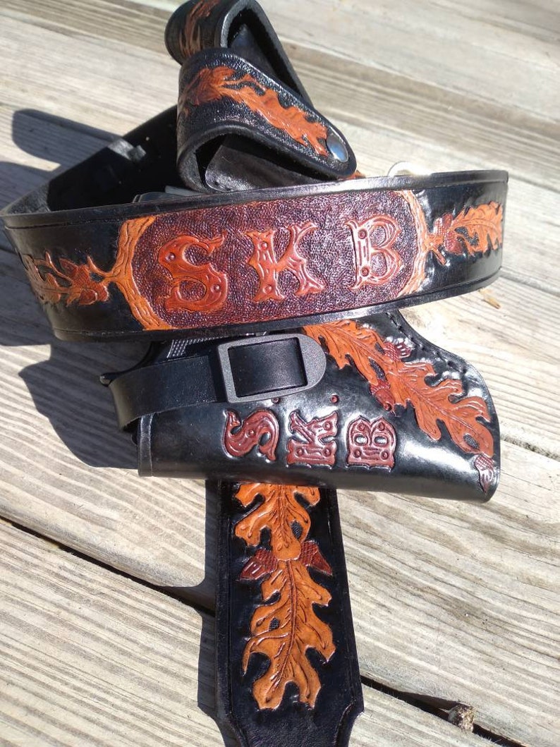 May include: A black leather belt and holster set with brown oak leaf and scrollwork designs. The belt features the initials "SKB" in a decorative font. The set is made of thick leather and has a buckle closure.