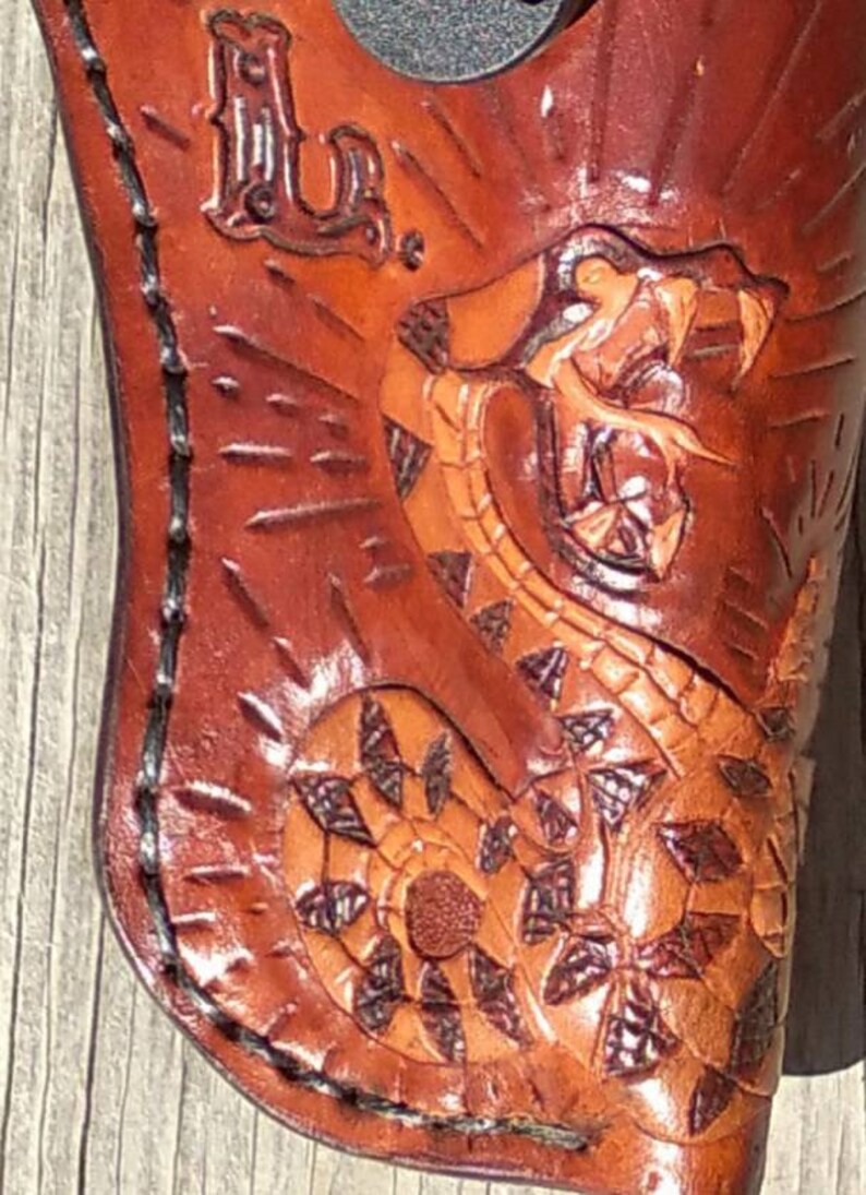 K&ouml;nnte beinhalten: Ein braunes Lederholster mit einem detaillierten Schlangendesign. Die Schlange ist in Orange- und Braunt&ouml;nen geschnitzt und gef&auml;rbt. Die Initialen "L.U." sind oben eingepr&auml;gt. Das Holster ist mit schwarzem Faden gen&auml;ht.