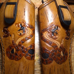 May include: Two brown leather holsters with a detailed snake design carved into the leather. Each holster has a black buckle and strap. The snake is depicted with its mouth open, revealing its fangs. The holsters are likely for a firearm.