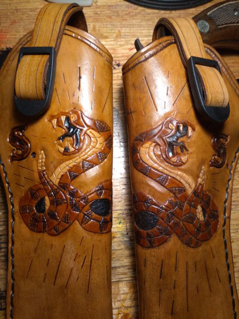 May include: Two brown leather holsters with a detailed snake design carved into the leather. Each holster has a black buckle and strap. The snake is depicted with its mouth open, revealing its fangs. The holsters are likely for a firearm.
