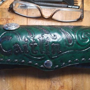 May include: Green leather eyeglass case with the name "Caitlin" embossed on the front. The case features decorative scrollwork and a silver-colored button closure. A silver-colored decorative button is on the top of the case.