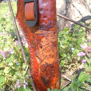 K&ouml;nnte beinhalten: Ein handgefertigter, brauner Lederholster mit einem detaillierten gepr&auml;gten Design eines Hasen und Bl&auml;ttern. Der Holster hat einen Lederriemen mit einer schwarzen Schnalle. Der Artikel befindet sich vor einem nat&uuml;rlichen Hintergrund im Freien.