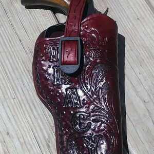 May include: A burgundy leather holster with a decorative floral design and the text "TAYLOR". The holster is designed to hold a revolver, with a strap and buckle closure. The revolver has a wooden grip.
