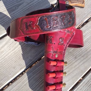 May include: A red leather gun holster and belt set with the text "K.S. 1875" and barbed wire detailing. The holster holds a revolver and several red shotgun shells.