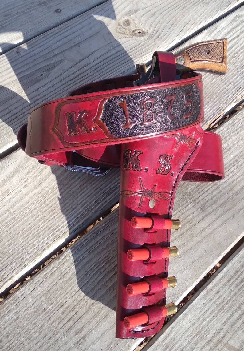 May include: A red leather gun holster and belt set with the text "K.S. 1875" and barbed wire detailing. The holster holds a revolver and several red shotgun shells.