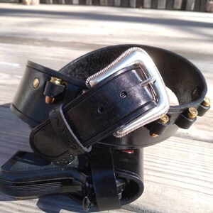 May include: Black leather gun holster with a silver buckle and decorative stitching. The holster is designed to hold a handgun and includes loops for ammunition. The belt is shown on a wooden surface.
