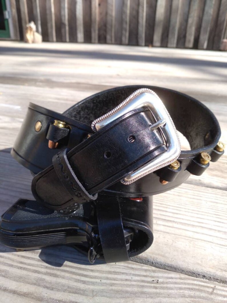 May include: Black leather gun holster with a silver buckle and decorative stitching. The holster is designed to hold a handgun and includes loops for ammunition. The belt is shown on a wooden surface.