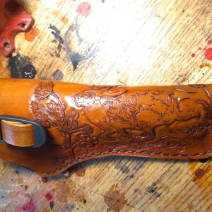 K&ouml;nnte beinhalten: Ein handgefertigtes, hellbraunes Lederholster mit einem detaillierten gepr&auml;gten Design mit einem Hasen und Bl&auml;ttern. Das Holster hat eine schwarze Schnalle und einen Riemen und ist mit schwarzem Faden gen&auml;ht. Der Artikel ist f&uuml;r eine Schusswaffe bestimmt.