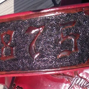 May include: Close-up of a red leather belt with embossed details. The belt features the year "1875" in a central, textured black panel, framed by decorative borders. The edges are a darker shade of red.