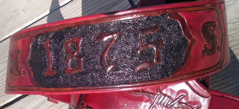 May include: Close-up of a red leather belt with embossed details. The belt features the year "1875" in a central, textured black panel, framed by decorative borders. The edges are a darker shade of red.