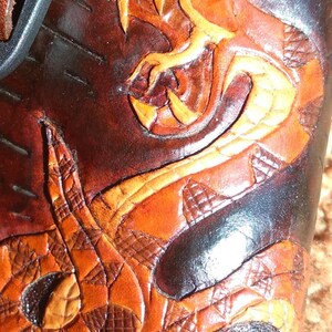 May include: Close-up of a brown leather item with a detailed snake design. The snake is carved and colored in shades of orange and yellow, with a textured pattern. The background is a darker brown.