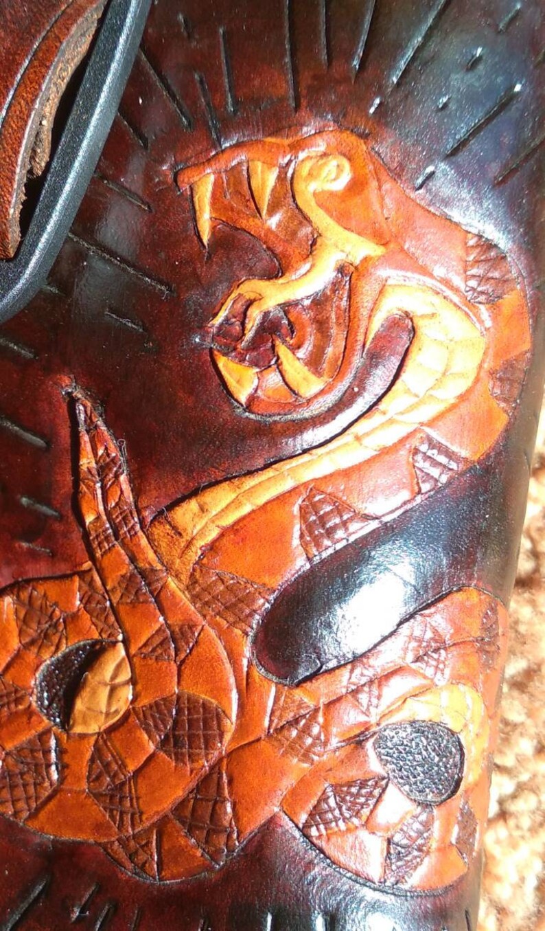 May include: Close-up of a brown leather item with a detailed snake design. The snake is carved and colored in shades of orange and yellow, with a textured pattern. The background is a darker brown.