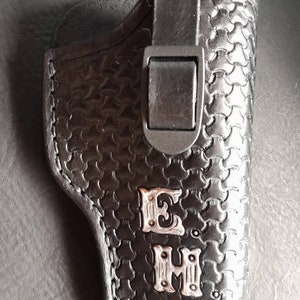 May include: Black leather holster with a textured pattern and a black clasp. The holster features silver-colored initials "E H". The design is a combination of functionality and style, suitable for carrying a firearm.