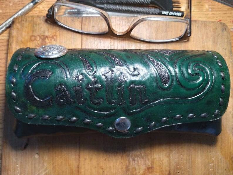 K&ouml;nnte beinhalten: Ein handgefertigtes, gr&uuml;nes Lederetui f&uuml;r Brillen mit dem Namen "Caitlin" auf der Vorderseite. Das Etui hat dekorative Pr&auml;gungen und einen silberfarbenen Knopfverschluss. Es dient zum Schutz von Brillen.