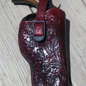 K&ouml;nnte beinhalten: Ein burgunderfarbenes Lederholster mit einem dekorativen Blumendesign und dem Text "TAYLOR". Das Holster ist f&uuml;r einen Revolver konzipiert, mit Riemen und Schnallenverschluss. Der Revolver hat einen Holzgriff.