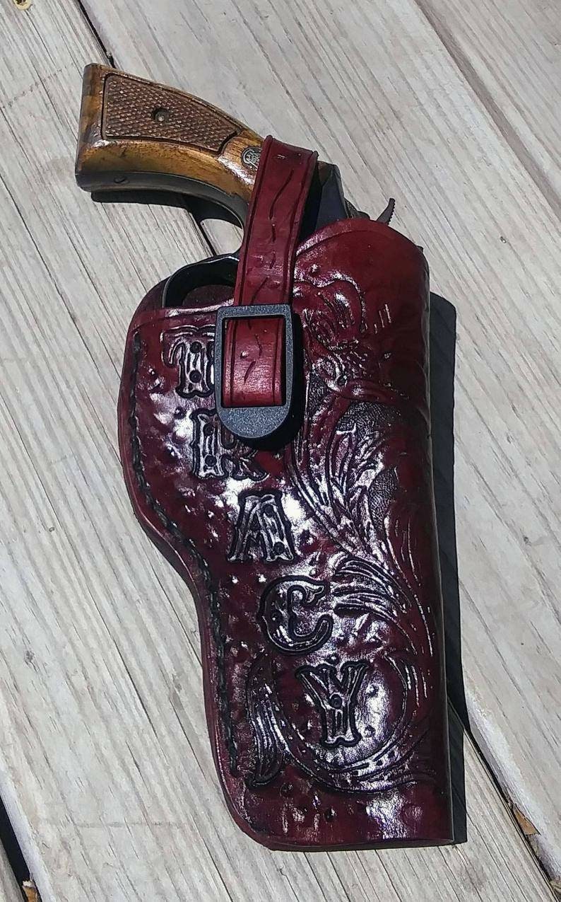 K&ouml;nnte beinhalten: Ein burgunderfarbenes Lederholster mit einem dekorativen Blumendesign und dem Text "TAYLOR". Das Holster ist f&uuml;r einen Revolver konzipiert, mit Riemen und Schnallenverschluss. Der Revolver hat einen Holzgriff.