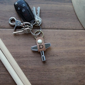 May include: A set of wooden drumsticks, a car key, and a leather key chain with a metal drum key. The key chain has the letter "D" engraved on it. The items are on a wooden surface.