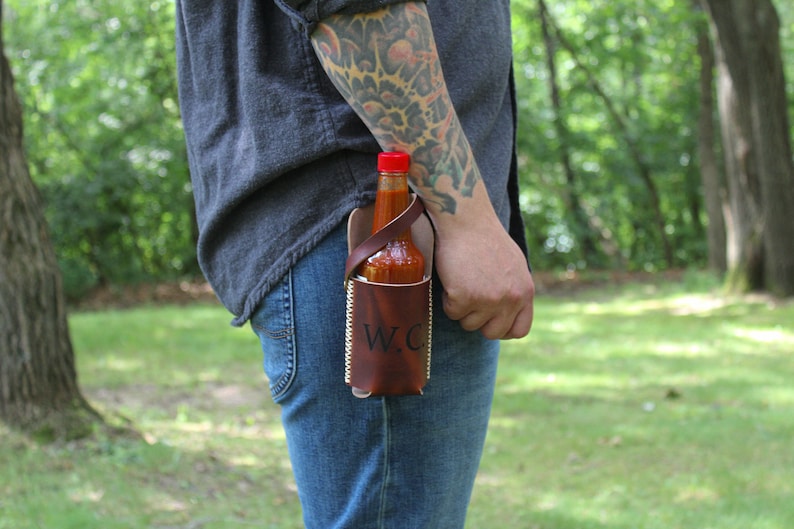 Peut inclure: Un porte-sauce piquant en cuir marron avec une sangle et les initiales "W.C." grav&eacute;es sur le devant. Le porte-sauce est attach&eacute; &agrave; la ceinture d'une personne et contient une bouteille en verre de sauce piquante.