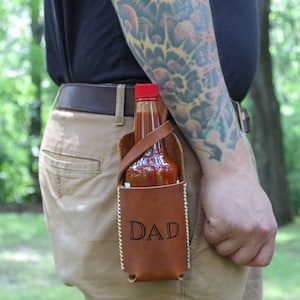 Peut inclure: Un porte-sauce piquant en cuir marron avec une sangle et le mot "DAD" grav&eacute; sur le devant. Une bouteille de sauce piquante est &agrave; l'int&eacute;rieur du porte-sauce.