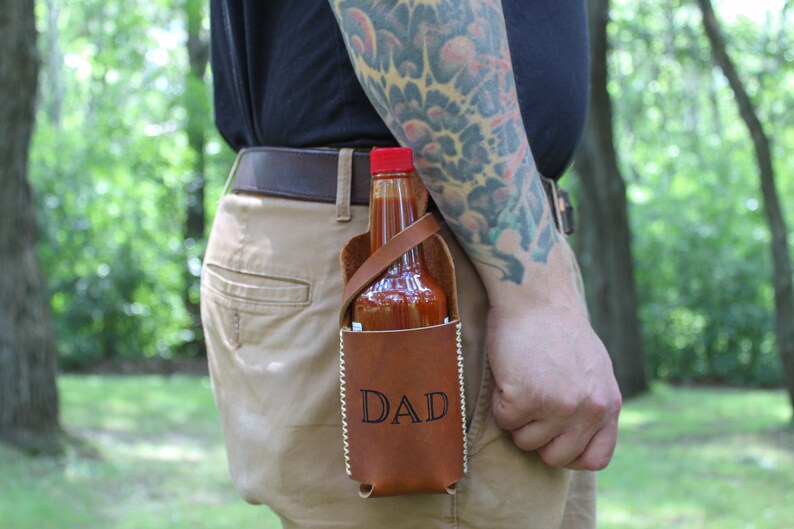 Peut inclure: Un porte-sauce piquant en cuir marron avec une sangle et le mot "DAD" grav&eacute; sur le devant. Une bouteille de sauce piquante est &agrave; l'int&eacute;rieur du porte-sauce.