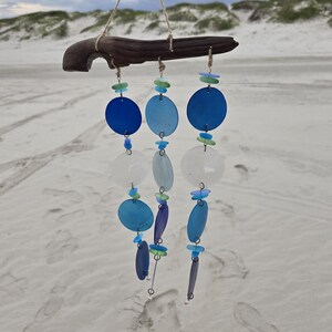 Peut inclure: Un carillon éolien artisanal avec des disques de verre bleus et blancs suspendus à un morceau de bois flotté. Le carillon est suspendu à un crochet métallique avec de la ficelle naturelle. Le fond est une plage de sable sous un ciel nuageux.