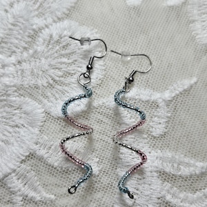 May include: A pair of handmade earrings with a spiral design. The earrings feature a silver-tone metal wire base, with alternating sections of light blue and pink beads. The earrings hang from silver-tone ear wires, and are displayed on a white lace surface.