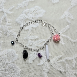 May include: A silver-toned chain bracelet with various charms. Charms include a black gemstone, a white elongated bead, a cluster of purple beads, a pink beaded ball, and a dark blue bead. The bracelet is displayed on a white lace surface.