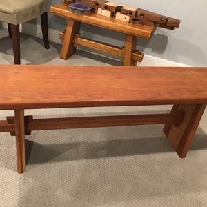 Puede incluir: Un banco de madera con un diseño simple y moderno. El banco tiene una parte superior rectangular y una sola viga de soporte horizontal. Las patas están hechas de madera maciza y tienen un diseño único y angulado.