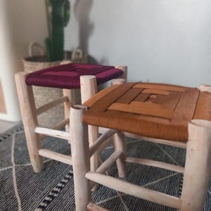 Op de afbeelding: Twee houten krukjes met gevlochten zittingen. Het krukje links heeft een bordeauxrode zitting en het krukje rechts heeft een bruine zitting.