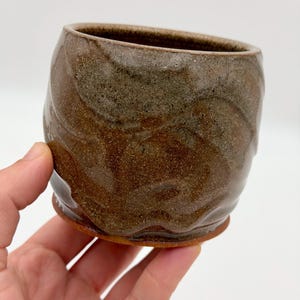 May include: A small, rounded ceramic cup featuring a mottled, earthy glaze with swirling patterns of dark brown, tan, and grey. The surface has a textured, organic appearance with a smooth rim and a slightly flared base, showcasing a handcrafted finish.