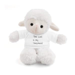 Puede incluir: Un juguete de peluche de cordero blanco con un cuerpo esponjoso, orejas pequeñas y ojos negros. El cordero lleva una camiseta blanca con el texto "The Lord is my Shepherd" en escritura negra. El juguete está diseñado para ser suave y tierno.