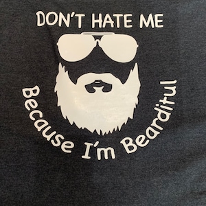 Puede incluir: Una camiseta gris oscuro con un diseño gráfico blanco. El gráfico presenta a un hombre de dibujos animados con gafas de sol y barba. El texto dice "No me odies porque soy barbudo."