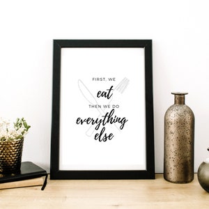 以下が含まれることがあります： 黒いフレームに入ったプリントで、黒い筆記体で「FIRST, WE EAT THEN WE DO everything else」と書かれています。 プリントは木製の表面に置かれています。 装飾的な花瓶と小さな植物も見えます。