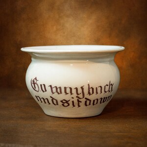May include: A white ceramic bowl with a flared rim and rounded body. The bowl features dark red gothic lettering that reads "Gomaybnch mdsitdow". The bowl is set against a brown background.