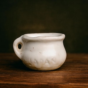 May include: A small, off-white ceramic cup with a handle. The cup has a rounded body and a slightly irregular rim. The word "JAPAN" is embossed on the side. The cup sits on a dark wooden surface.