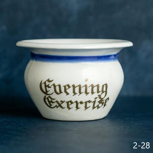 May include: A white ceramic pot with a blue stripe and the words "Evening Exercise" in gold lettering. The pot has a rounded body and a flared rim. The background is a dark blue.