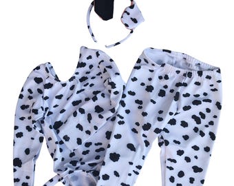 May include: A long-sleeved leotard and matching leggings set featuring a white fabric with a black cow-print pattern. The set includes a thin headband with two attached ears, one black and one white with a black spot, matching the cow theme.
