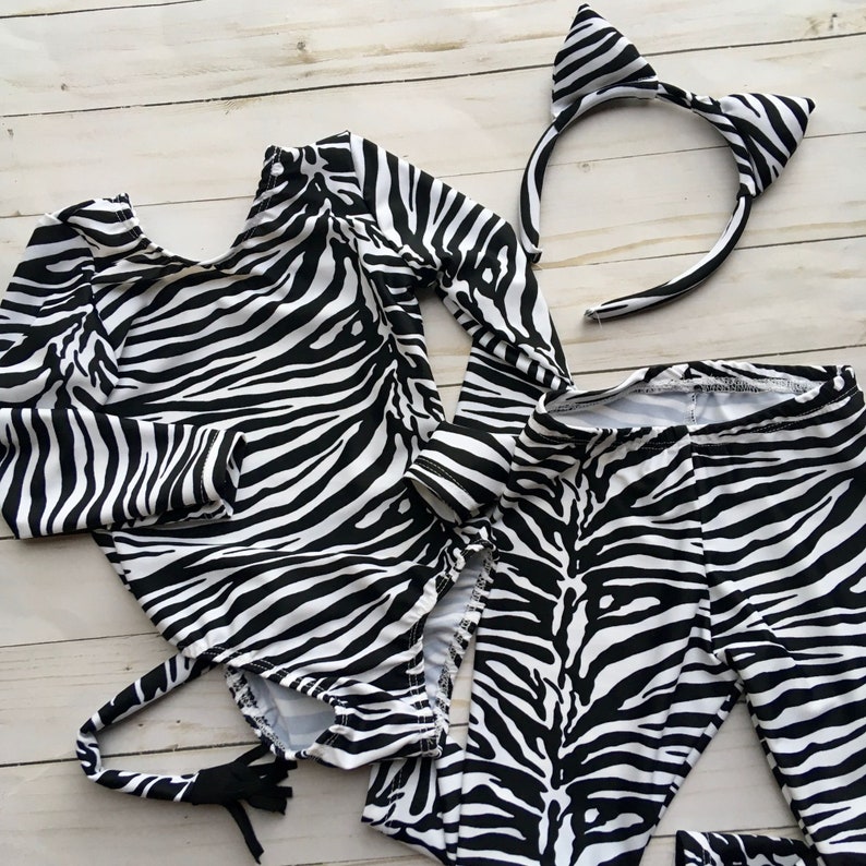 Może przedstawiać: Kostium zebry w czarno-białe pasy dla dziecka, w skład kt&oacute;rego wchodzi body z długim rękawem, legginsy, ogon i opaska na głowę z uszami.
