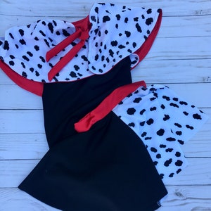 May include: A black dress featuring a white capelet with a black dalmatian spot pattern and a red tie closure at the neckline. The dress includes a matching red waistband and a skirt section with a white dalmatian spot pattern on the right side.