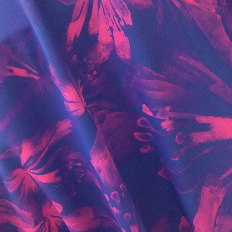 May include: Close-up of a fabric with a floral pattern. The design features large, stylized flowers in shades of pink and magenta against a deep blue background. The fabric is draped, creating soft folds and shadows that enhance the visual texture.