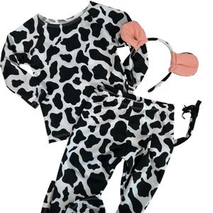 May include: A cow-themed costume set featuring a long-sleeved shirt and pants, both in white with black spots. The set includes a headband with pink ears and a black and white tail. Perfect for a costume party or dress-up.