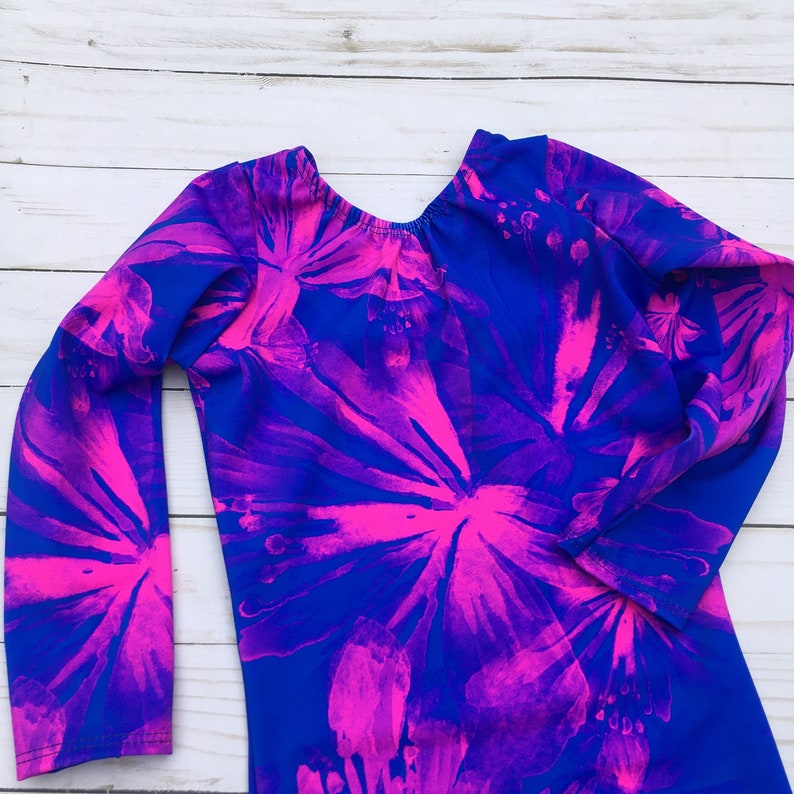 May include: A long-sleeved leotard with a vibrant floral pattern. The leotard features a royal blue base with large, bright pink flower designs. The garment is designed for gymnastics or dance, with a scoop back and a comfortable fit.