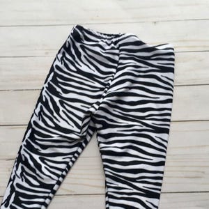 May include: Black and white zebra print leggings. The leggings feature a repeating pattern of black stripes on a white background. The fabric is smooth and appears to be stretchy. The waistband is elasticized. The leggings are shown on a light wood surface.