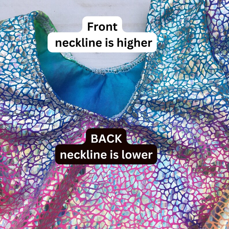 May include: Close-up of a leotard with a holographic iridescent finish. The front neckline is higher than the back neckline. The fabric has a textured, mosaic-like pattern.