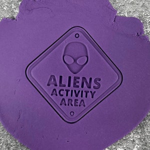 May include: Purple play dough with a black and white alien activity area sign pressed into it. The sign is a diamond shape with an alien head and the text "Aliens Activity Area".